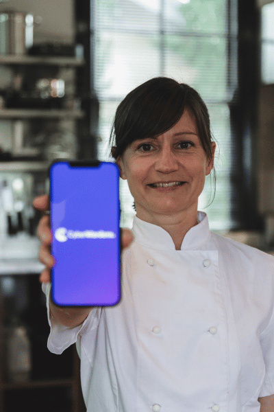 Female chocolatier in a chisp white chefs coat proudly holds her photo to the eye of the viewer displaying the Cyber Wardens mobile screen